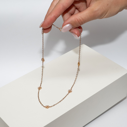 Hand holding a delicate thin flower necklace out of rose gold with diamonds over a white box on a light gray background